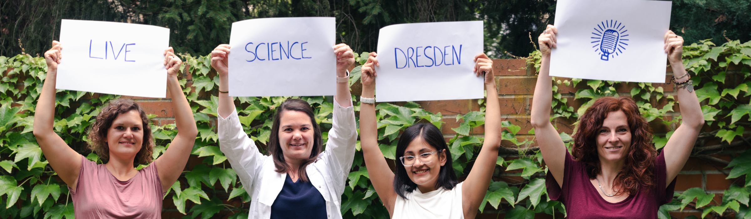 Das Team steht in einer Reihe vor einer Mauer und hält vier weiße Plakate über ihren Köpfen, auf denen Live Science Dresden steht.